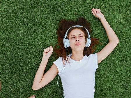 A woman lying on the green grass of the lawn listening to music in headphones and singing in a white T-shirt happinessの写真素材