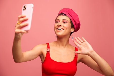 A young woman blogger with colored pink hair and a short haircut takes a picture of herself on the phone and broadcasts a smile in stylish clothes and a hat on a pink background monochrome styleの写真素材