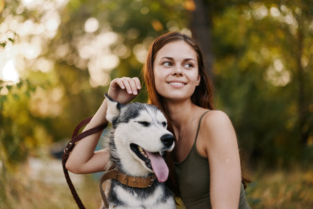 A woman with a husky breed dog smiles and affectionately strokes her beloved dog while walking in nature in the park in autumn against the backdrop of sunsetの写真素材