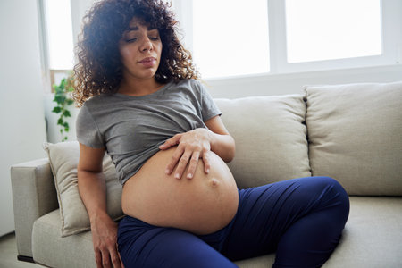 Pregnant woman sitting on the couch lower back pain and headache, strain on the spine during pregnancy. Lifestyle difficulties of motherhood preparation for childbirth, the last month of pregnancyの写真素材