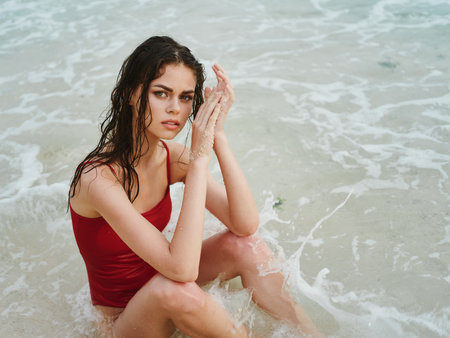 A woman with wet hair sits on the sand and looks at the camera, a slender tanned body on the ocean beach in Bali, the concept of traveling the world and vacationing in the islandsの写真素材