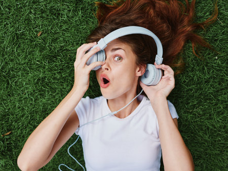 Woman in headphones listening to music smile with teeth lying on the green grass in summer, good mood spring surpriseの写真素材