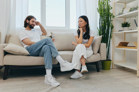 A man looks at his phone screen and smiles during an argument with his girlfriend. The angry and hurt woman looks in his direction and is sad. Family discord at home, phone addictionの写真素材