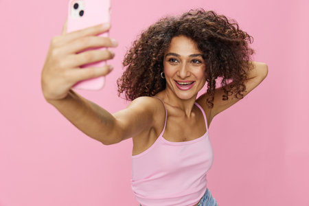 Woman blogger holding phone video call takes selfies, with curly hair in pink smile t-shirt and jeans poses on pink background, copy space, technology and social media, onlineの写真素材