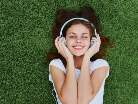 A happy woman lying on the green lawn in a white T-shirt and listening to music with her headphones smiling with her hands in upの写真素材