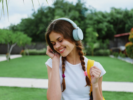 Happy teenage girl listening to music with headphones with a backpack in a white T-shirt and walking to college to study on the street smiling with teeth, student lifestyleの写真素材