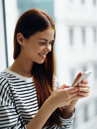 Woman holding her phone and looking and smiling at it near the window of the house, online shopping...