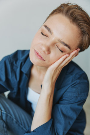 A young woman sitting in a chair at home smiling with teeth with a short haircut in jeans and a denim shirt on a white background. Girl natural poses with no filtersの写真素材