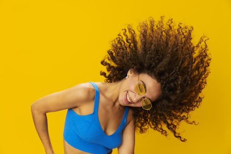 Woman with curly afro hair in a blue T-shirt on a yellow background dancing flying hair with sunglasses yellow, hand signs, look into the camera, smile with teeth and happiness, copy spaceの写真素材