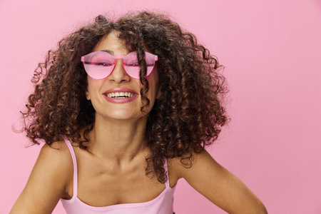 Woman with curly afro hair with sunglasses model poses on a pink background in a pink T-shirt, free movement and dance, look into the camera, smile with teeth and happiness, copy spaceの写真素材