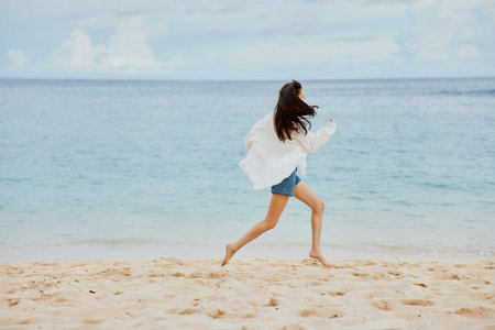 Sports woman runs along the beach in summer clothes on the sand in a yellow T-shirt and denim shorts white shirt flying hair ocean viewの写真素材