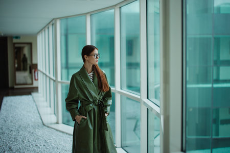 Fashion woman in a stylish raincoat and sunglasses standing by the window and looking at the city and the streetの写真素材