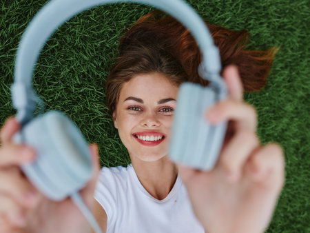 A girl lies on the grass and holds out her headphones to the camera smiling with her teethの写真素材