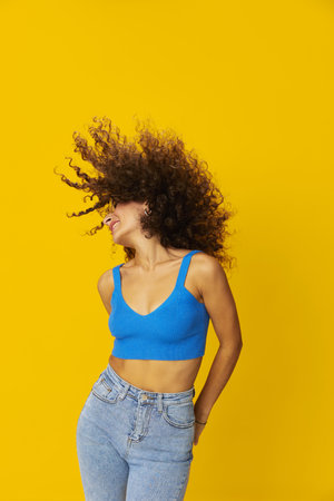 Woman with curly afro hair in a blue T-shirt on a yellow background dancing flying hair with sunglasses yellow, hand signs, look into the camera, smile with teeth and happiness, copy spaceの写真素材