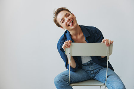 A young woman sitting in a chair at home smiling with teeth with a short haircut in jeans and a denim shirt on a white background. Girl natural poses with no filtersの写真素材
