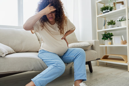 Pregnant woman sitting on the couch at home and wearing pregnancy-friendly shoes with a problem due to the stomach, headache, pregnancy and motherhood difficulties, severe fatigue, headacheの写真素材