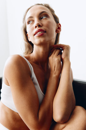 Portrait of a woman with a beautiful tanned body posing on the couch in white lingerie on a white background at home. Taking care of body and skin healthの写真素材