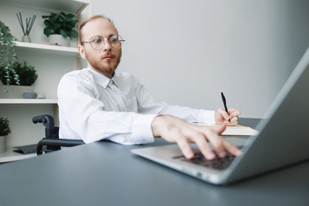 A man wheelchair businessman in the office works at a laptop, writing down ideas and a plan in a...