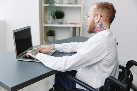 A man wheelchair businessman with tattoos office works at a laptop online, social networks and startup, integration into society, the concept of working a person with disabilities, view from the backの写真素材