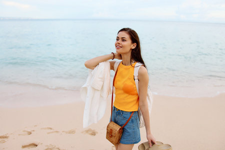 Woman smile with teeth after swimming in the ocean with a backpack in a wet yellow tank top and denim shorts walks along the beach, summer vacation on an island by the ocean in Bali sunsetの写真素材
