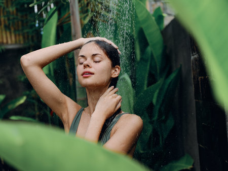 A woman takes a shower and washes her head and hair outdoors in nature, closed eyes and a smile on the background of tropical plants, palm trees, green banana leaves, summer rain, lifestyleの写真素材