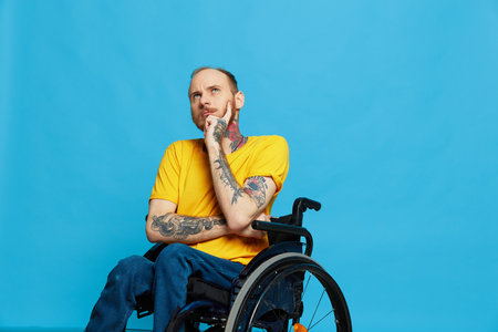a man sits in a wheelchair thoughtfulness in a t-shirt on a blue background in the studio, the...