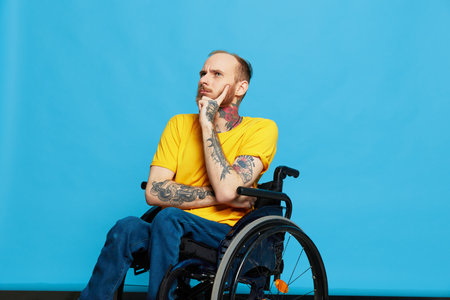 a man sits in a wheelchair thoughtfulness in a t-shirt on a blue background in the studio, the concept of a free barrier-free environment for people with disabilitiesの写真素材