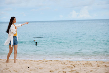 Sports woman runs along the beach in summer clothes on the sand in a yellow tank top and denim shorts white shirt flying hair ocean view, beach vacation and travelの写真素材