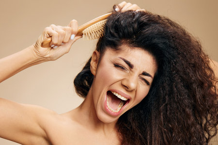 LONG HAIR PROBLEMS. NAUGHTY HAIR CONCEPT. Suffering screaming angry tanned curly Latin lady with hairbrush suffer with unruly hair posing isolated on pastel beige background, close eyes. Copy spaceの写真素材
