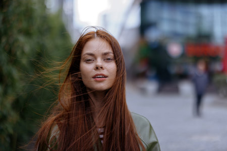 Portrait of a beautiful young girl in the city looking into the camera with red flying hair in a green raincoat in the city against a background of bamboo in spring, lifestyle in the cityの写真素材