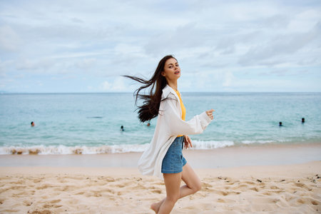Sports woman runs along the beach in summer clothes on the sand in a yellow tank top and denim shorts white shirt flying hair ocean view, beach vacation and travel freedomの写真素材