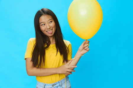 Asian woman holding a yellow balloon on a blue background in a yellow T-shirt smiling with teeth, funny, crooked and showing her tongueの写真素材