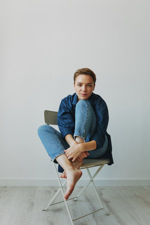A young woman sitting in a chair at home smiling with teeth with a short haircut in jeans and a denim shirt on a white background. Girl natural poses with no filtersの写真素材