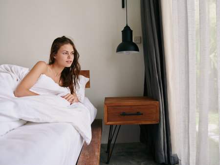 A woman after a shower with wet hair lies on a bed under a blanket in a bedroom near a window, a healthy sleep on an orthopedic mattress and a memory foam pillow, body and hair careの写真素材
