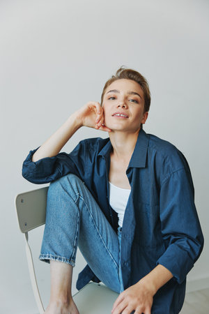 A young woman sitting in a chair at home smiling with teeth with a short haircut in jeans and a denim shirt on a white background. Girl natural poses with no filtersの写真素材