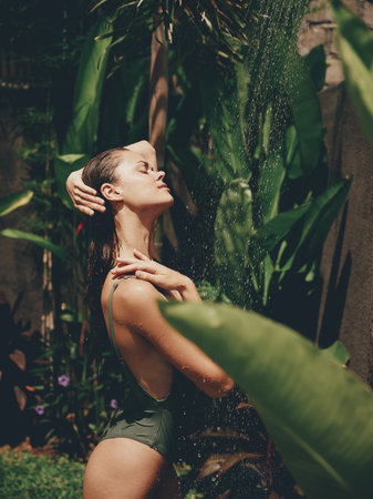 Sexy woman, body in a swimsuit takes a tropical shower outdoors against the backdrop of green tropical leaves, flowers and palm trees. Summer outdoor recreation, tanned skinの写真素材