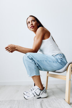 A woman sitting on a full-length chair in a studio against a white wall with very tanned skin in blue jeans and a white T-shirtの写真素材