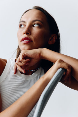 A young beautiful woman sits on a chair in stylish clothes and poses in fashionable style against a...