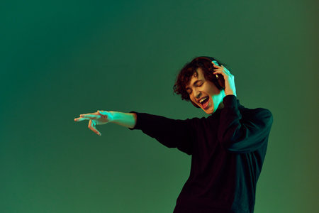 Man teenager wearing headphones listening to music, dancing and singing with his eyes closed, DJ party happiness and smile, hipster lifestyle, portrait green background mixed neon light, copy spaceの写真素材