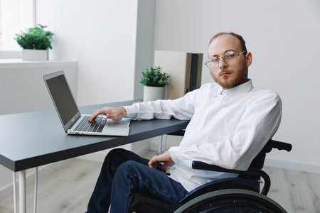 A man in a wheelchair looking at the camera businessman in the office working on a laptop online,...