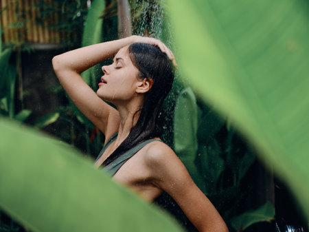 A woman in a swimsuit washes her head in a tropical shower outdoors against the backdrop of green tropical leaves, flowers and palm trees. Body and hair care, tanned skin, sexy, open mouthの写真素材