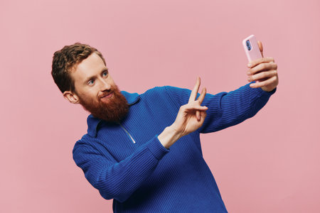 Portrait of a redheaded man with phone in hand taking selfies and photos on his phone with a smile on a pink background, bloggerの写真素材