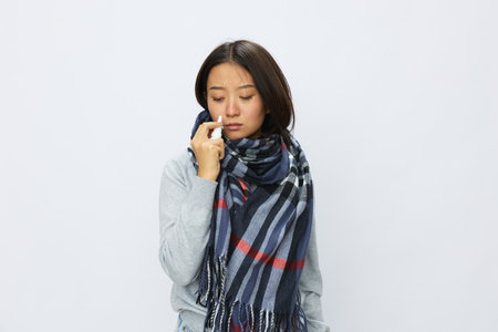 Asian woman flu cold with stuffy nose holds nasal spray and sprays nasal spray for allergies and covid 19 virus with sore throat on white backgroundの写真素材