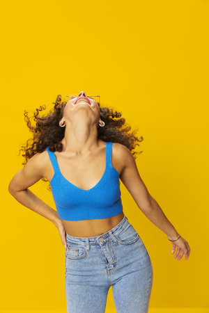 Woman with curly afro hair in a blue T-shirt on a yellow background dancing flying hair with...