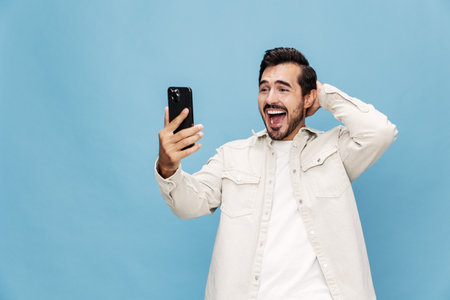 Portrait of a brunette man with a phone in his hands, a smile with teeth of surprise and happiness, on a blue background in a white T-shirt and jeans, copy spaceの写真素材