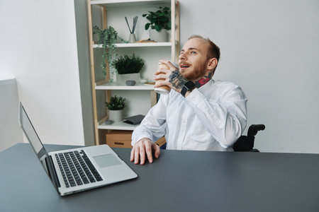 A man in a wheelchair, a businessman with tattoos in the office at a laptop, a coffee break, the concept of working a person with disabilities, freedom from social framesの写真素材