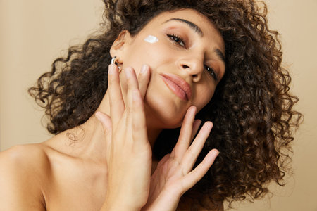 Woman beauty face close-up applying anti-aging moisturizer with fingers of her hand, skin health nails and hair, hair dryer style curly afro hair, body and beauty care conceptの写真素材