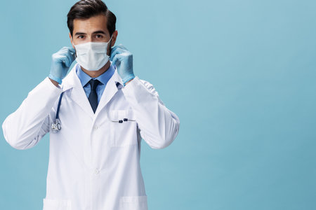 Male doctor in a white coat and medical mask looks at the camera on a blue isolated background, copy space, space for textの写真素材