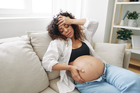 Pregnant woman headache lies at home in a shirt and jeans on the couch fatigue and heaviness in the last month of pregnancy before childbirth, motherhood difficulties, nauseaの写真素材
