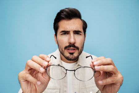 Close-up portrait of a brunette man looking through glasses that he holds in his hands, eye problems, glasses for vision farsightedness and nearsightedness, on a blue background, copy spaceの写真素材